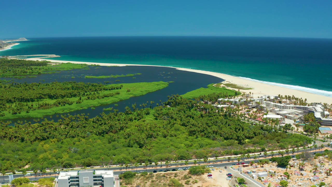 High aerial frame of San Jose Del Cabo coastline with dark lagoon water, bright green vegetation, white sand, deep blue ocean, small buildings, road activity, and no visible people.