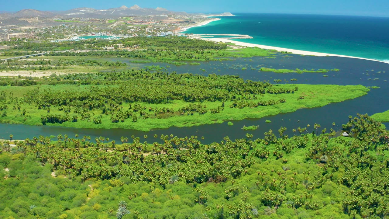 At 0:00, San Jose del Cabo wetlands fill the foreground below dark river water, with a pale beach, turquoise ocean, distant town, low mountains, and no visible people.