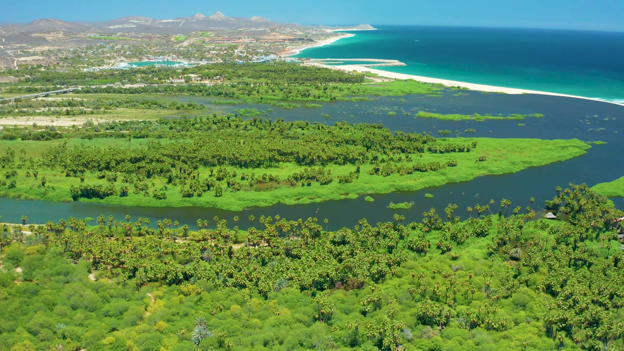 At 0:02, the frame shows a broad green estuary crossed by dark water, with a long sandy beach and deep blue ocean stretching along San Jose del Cabo, and no visible people.