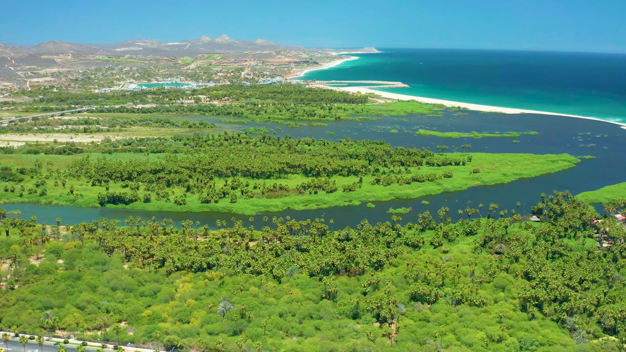 Aerial coastal landscape with river channels, dense greenery, road, beach, ocean, and hills.