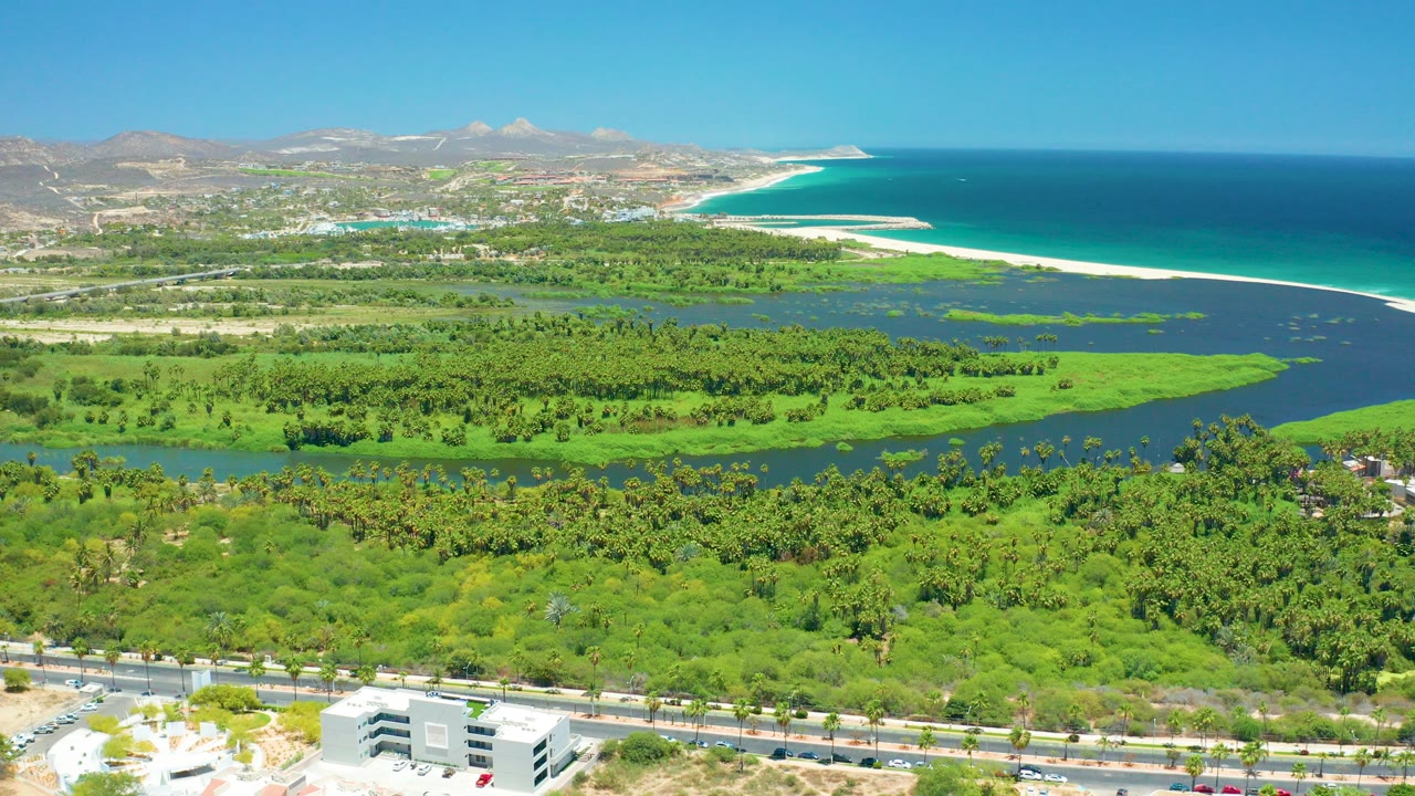 Wide aerial frame over San Jose del Cabo showing green wetland channels, a road and buildings in the foreground, blue ocean and sandy beach in the distance, and no visible people.