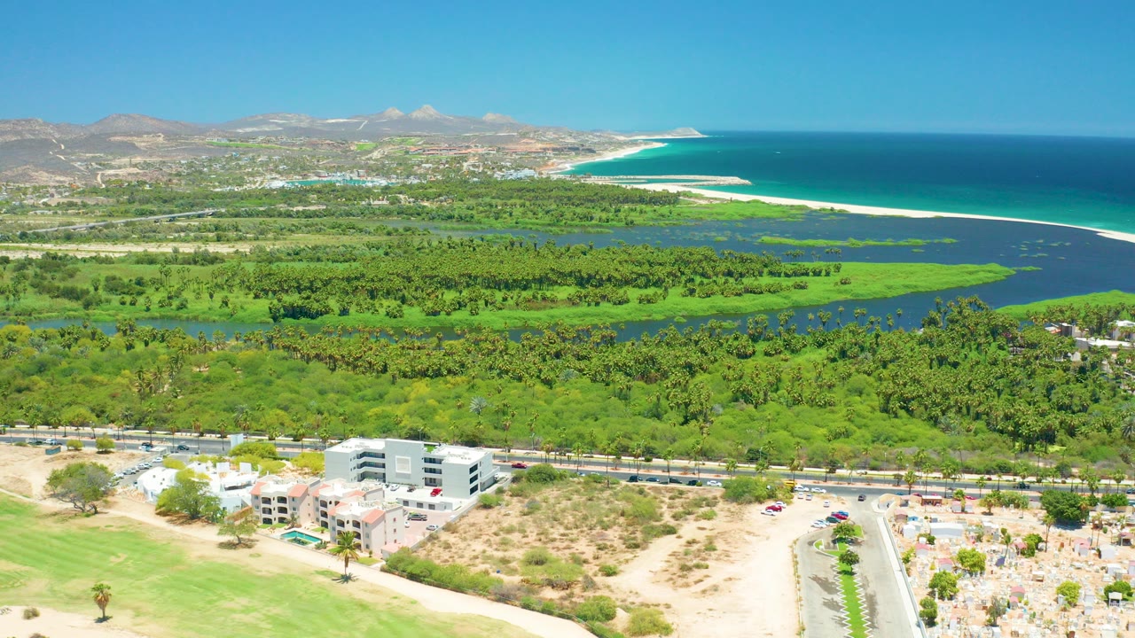 Opening aerial frame over San Jose del Cabo showing foreground roads and buildings, a green lagoon, a pale beach, blue ocean, distant mountains, and no visible people.