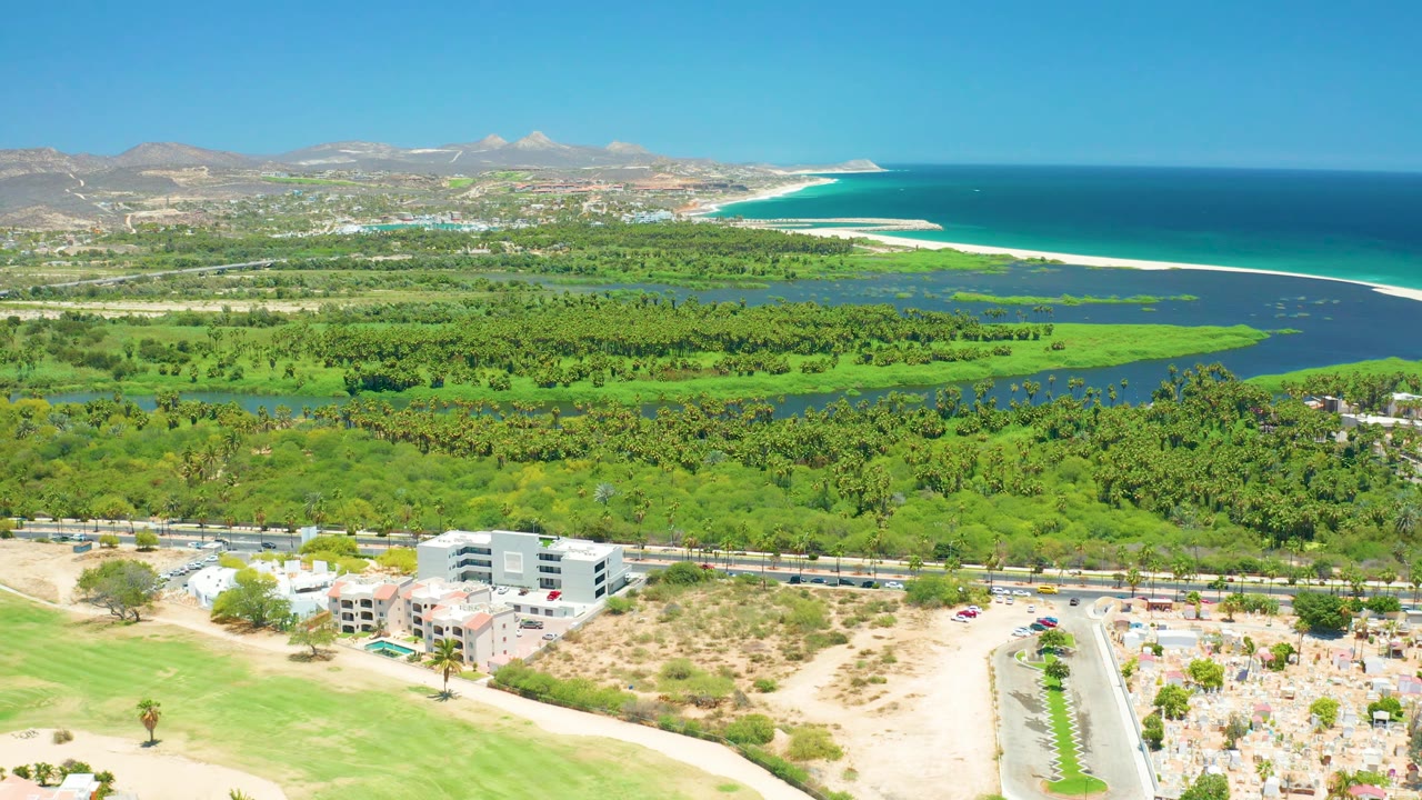 At two seconds, the San Jose del Cabo coastline stretches across the background while the lagoon, tree canopy, roads, modern buildings, cars, and cemetery area are visible in bright sun with no visible people.