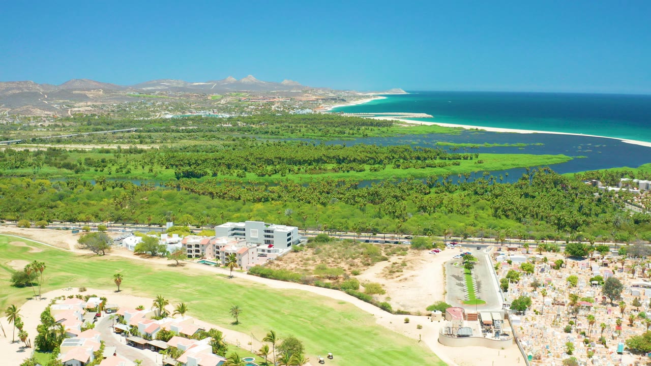 Wide aerial view over San Jose Del Cabo with green grounds and beach structures in the foreground, resort buildings, a sandy beach with umbrellas, blue ocean, and distant mountains.