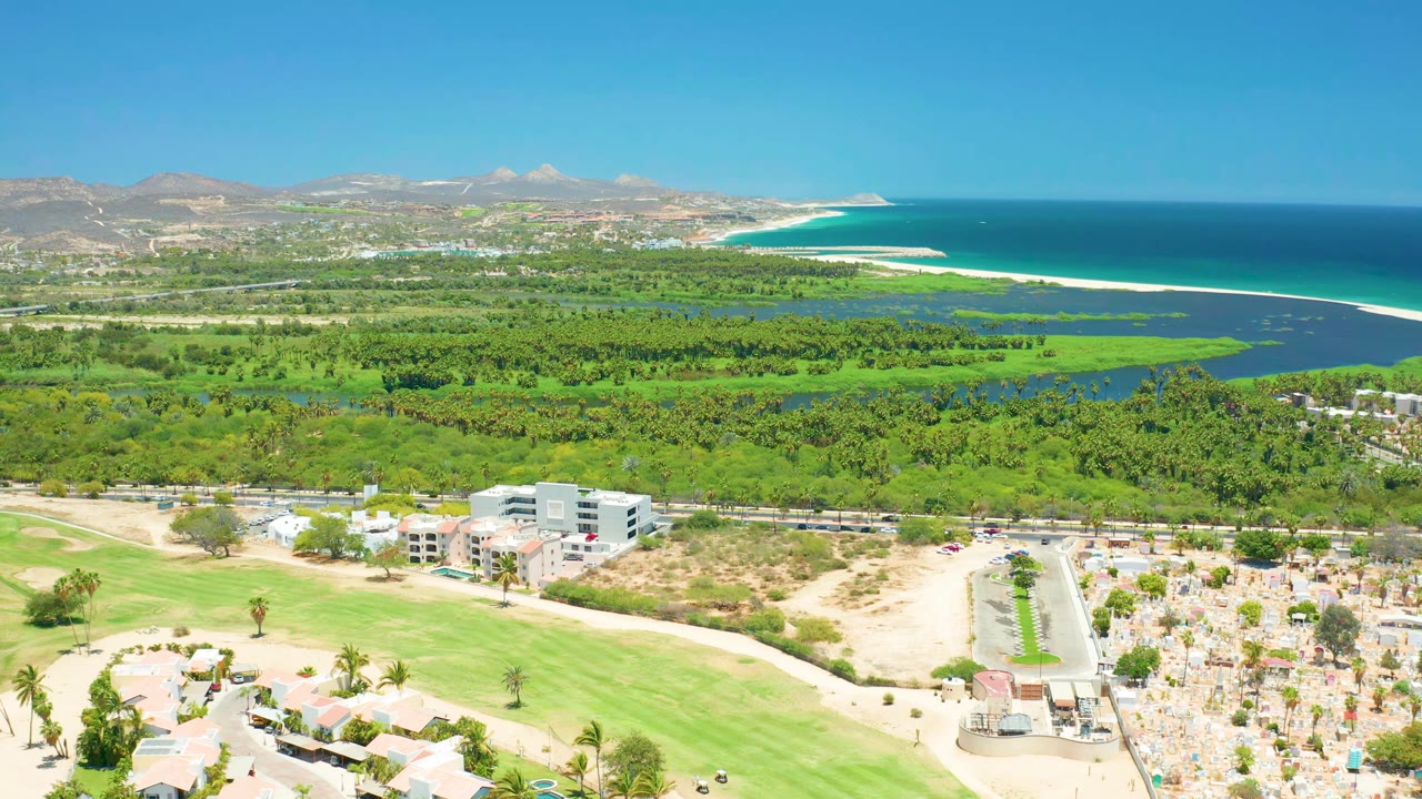 High drone view of coastal greenery, beach umbrellas, ocean, and mountains.