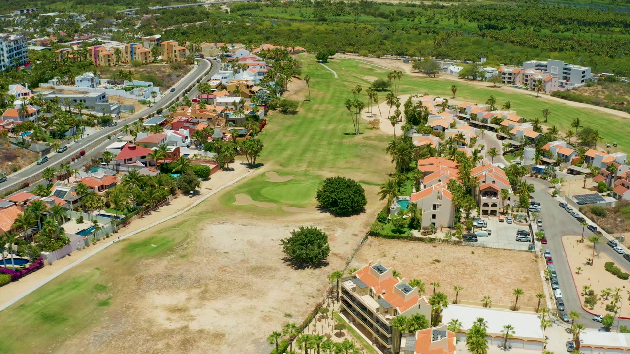 Opening aerial frame over San Jose Del Cabo with golf course fairways, sand traps, a curving highway, red-roof residential buildings, dry lots, palm trees, background greenery, and no visible people.