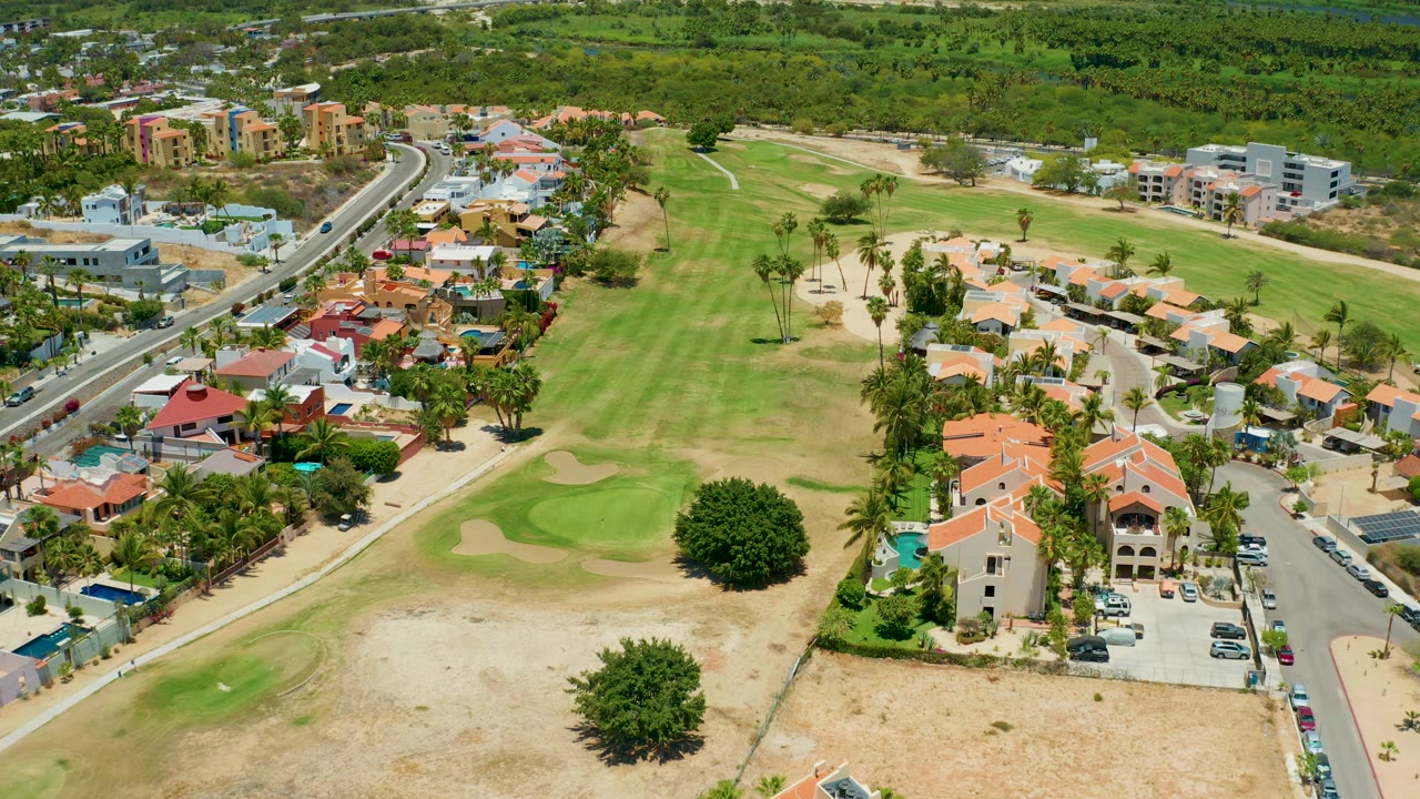 Aerial frame over San Jose Del Cabo with fairway grass and bunkers on the left, residential buildings and a road corridor on the right, and no visible people.