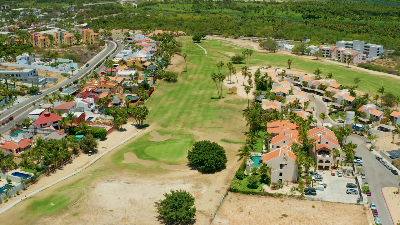 High aerial view in San Jose Del Cabo showing a curved road with vehicles, clusters of homes, fairway grass, sand traps, palm trees, and no visible people.