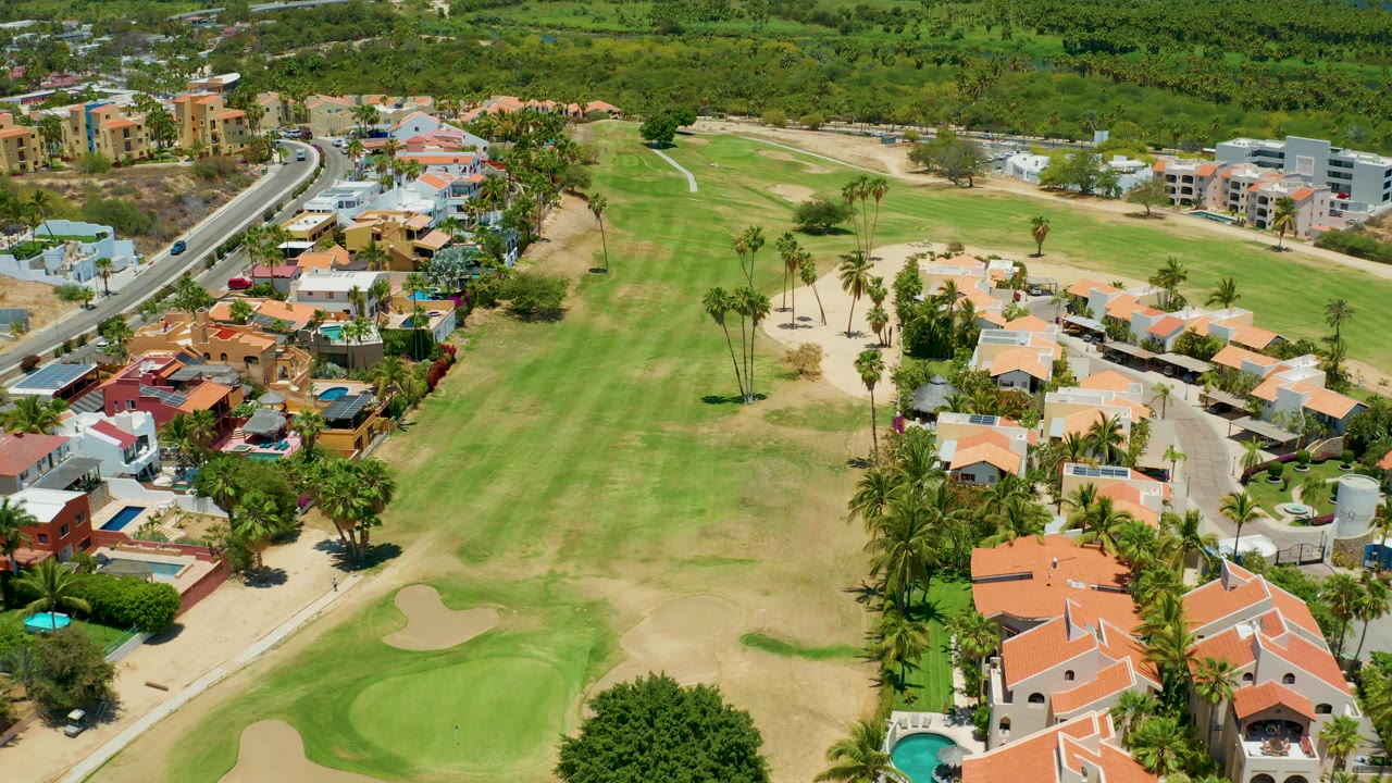 Aerial frame over San Jose Del Cabo with the fairway centered, homes and pools along the lower right, a road with cars on the left, and no visible people.