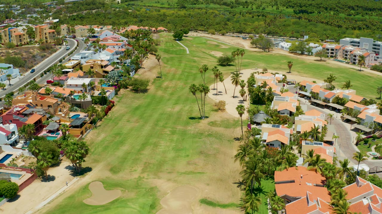 Golf course and neighborhood aerial with palms, houses, pools, and a road on the left.