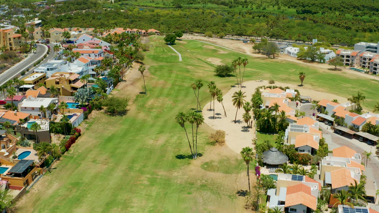 Aerial frame over San Jose Del Cabo showing orange-roofed homes, a green golf fairway, sand traps, palm trees, and surrounding vegetation, with no visible people.