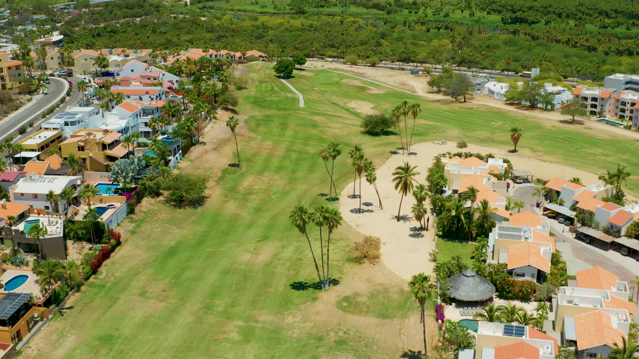 Aerial view across San Jose Del Cabo showing a broad green fairway, pale sand traps, a row of homes at the lower edge, and thick vegetation beyond the course, with no visible people.