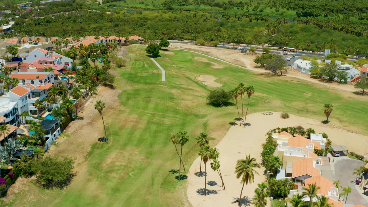Aerial view over a San Jose Del Cabo golf course with green fairway, tan sand areas, orange-roofed homes, palm trees, dense vegetation, and no visible people.