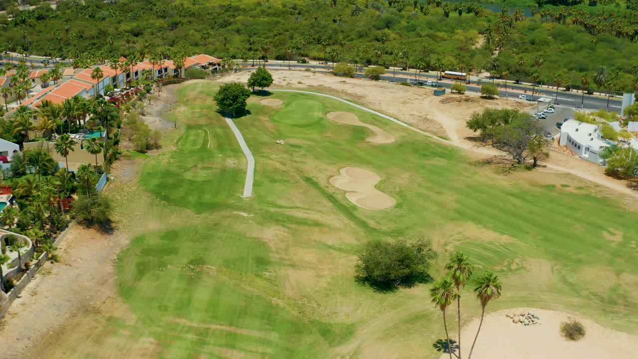 Opening aerial frame over a San Jose Del Cabo golf course, showing green and dry fairway patches, beige sand traps, a pale cart path, roadside buildings, trees, and no visible people.
