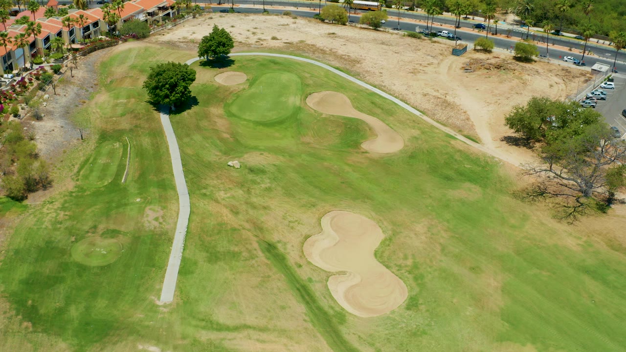 Aerial view over a San Jose Del Cabo golf course with green fairway, sand traps, a pale cart path, roadside dirt, buildings at the upper edge, and no visible people.