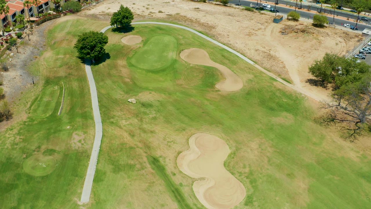 Drone view of golf fairway, pale cart path, sand traps, and a tree near the road.