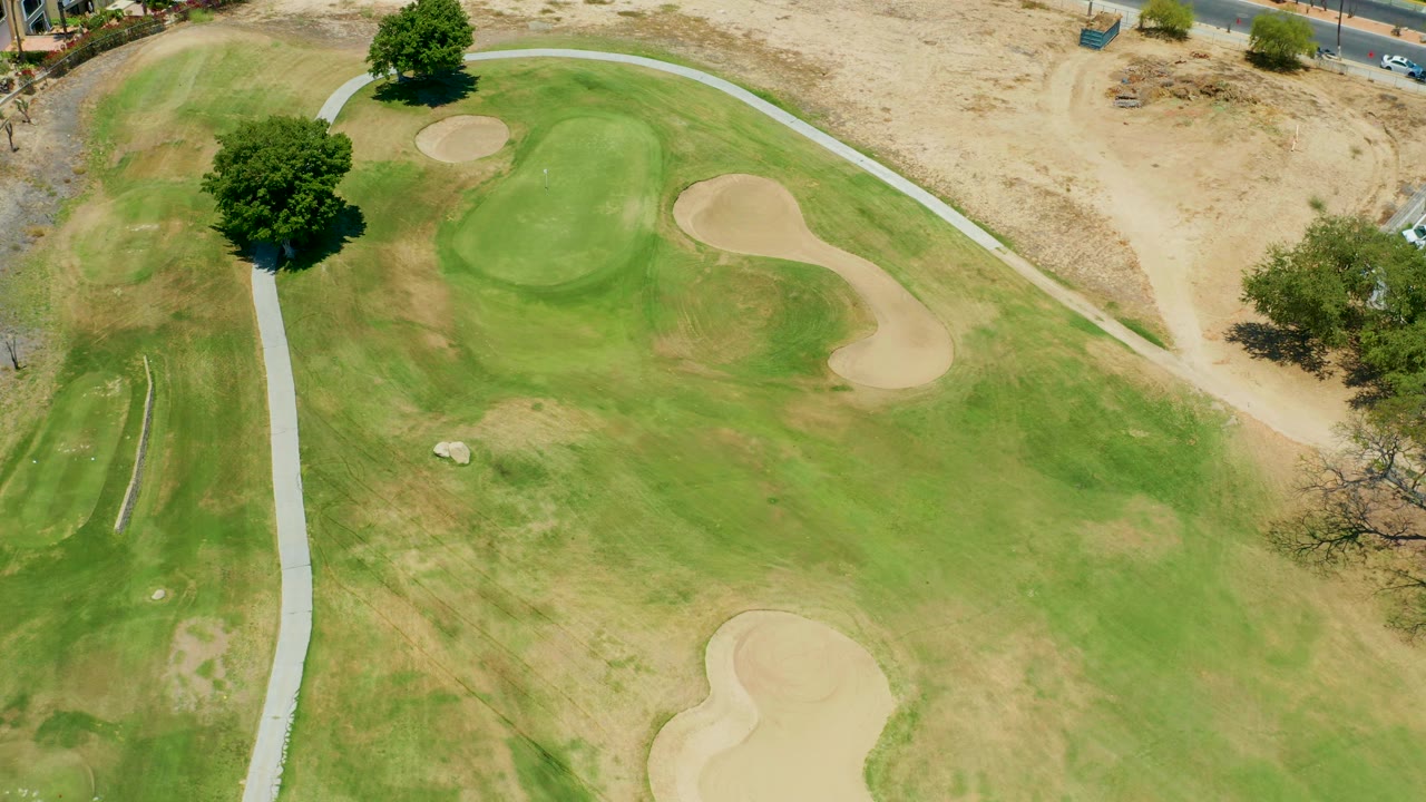Aerial frame over the San Jose Del Cabo course with the central green, tan bunkers, curved cart path, dry upper ground, trees casting shadows, and no visible people.