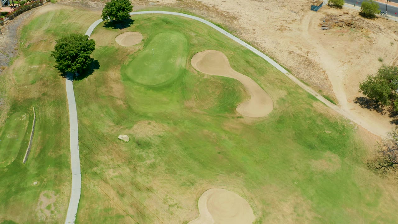 Overhead San Jose Del Cabo frame with the golf green, tan sand traps, a vertical pale cart path, dry dirt outside the course, tree shadows, and no visible people.