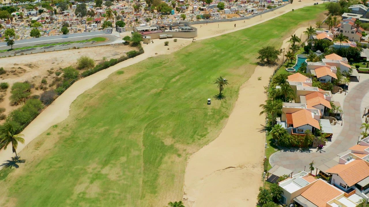 High-angle San Jose Del Cabo frame with the golf cart crossing the green, pale sand bands around the fairway, clustered homes to the right, road at the top, and no visible people.