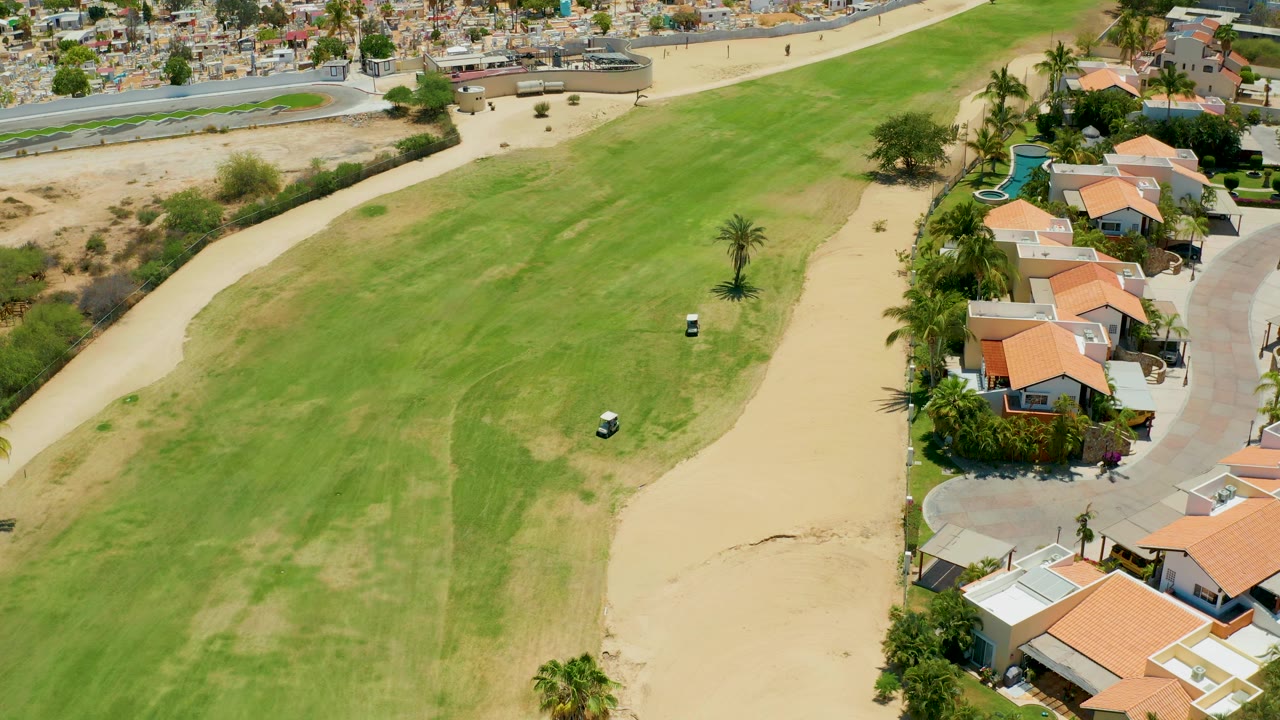 A high drone view in San Jose del Cabo shows the fairway stretching diagonally, a small golf cart near the center-right grass, sandy course edges, nearby houses, and no visible people.