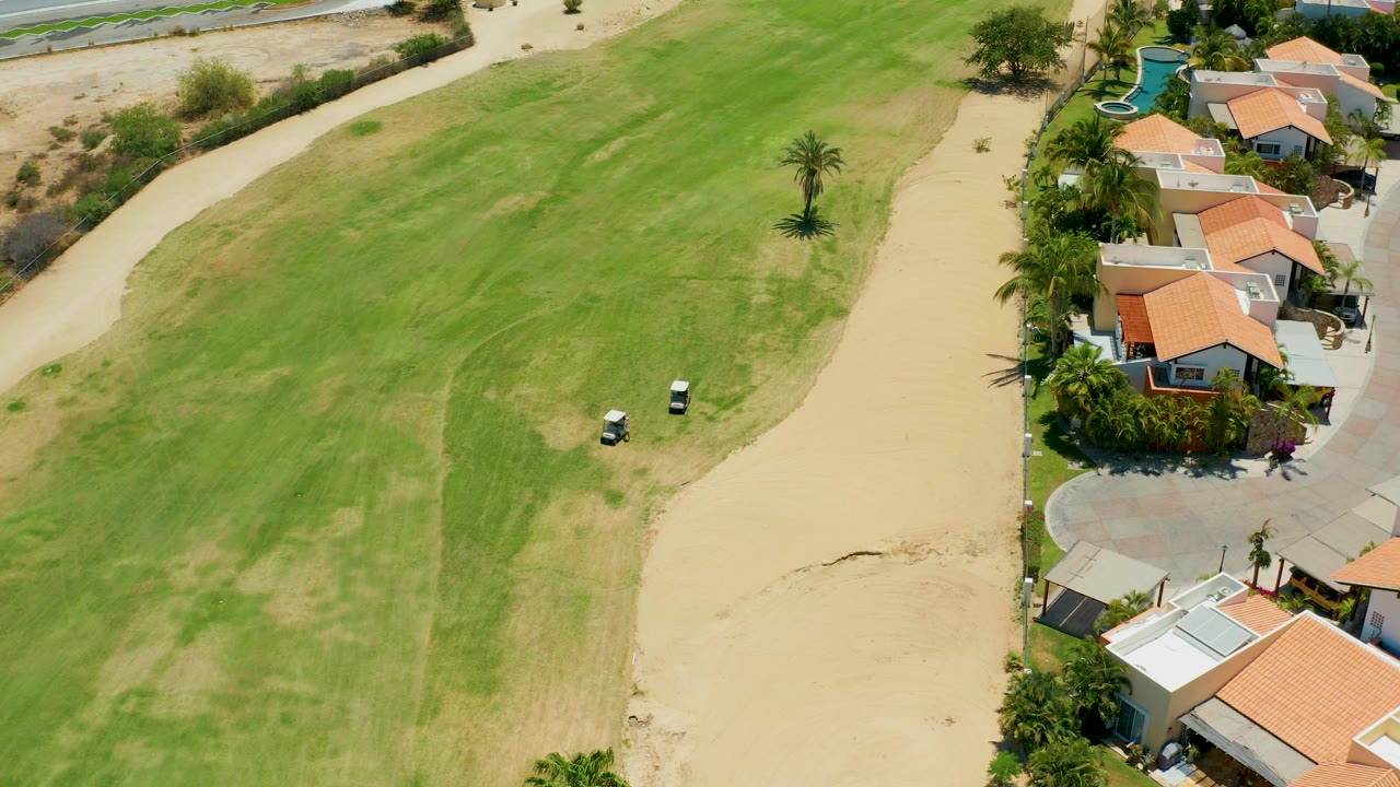 Overhead view in San Jose del Cabo of a green golf fairway, tan sand path, orange-roofed homes, palms, a pool, two white golf carts, and a small person near the residential edge.