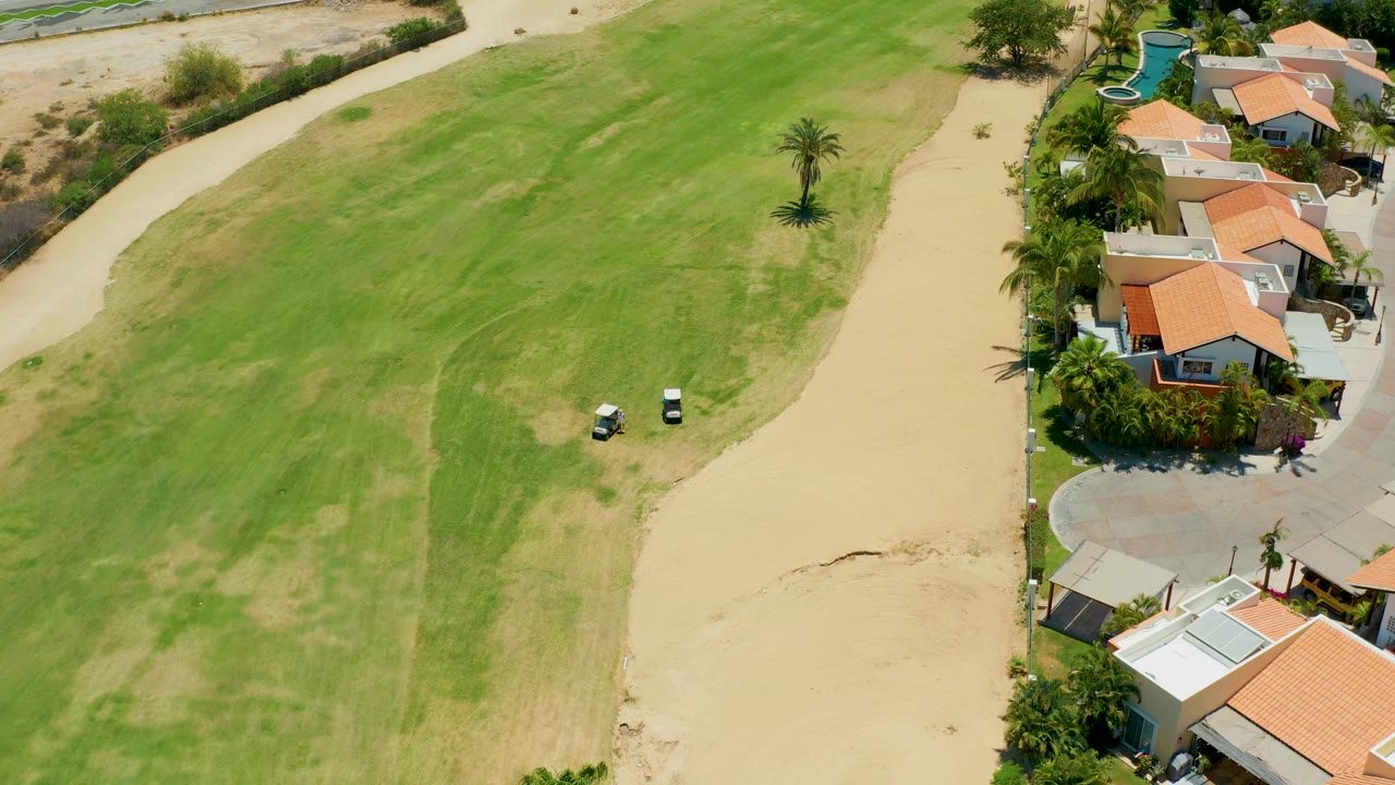 Overhead San Jose del Cabo view with no visible people, showing two white golf carts on the fairway, a broad tan path, scattered palms, and orange-roofed homes along the right edge.