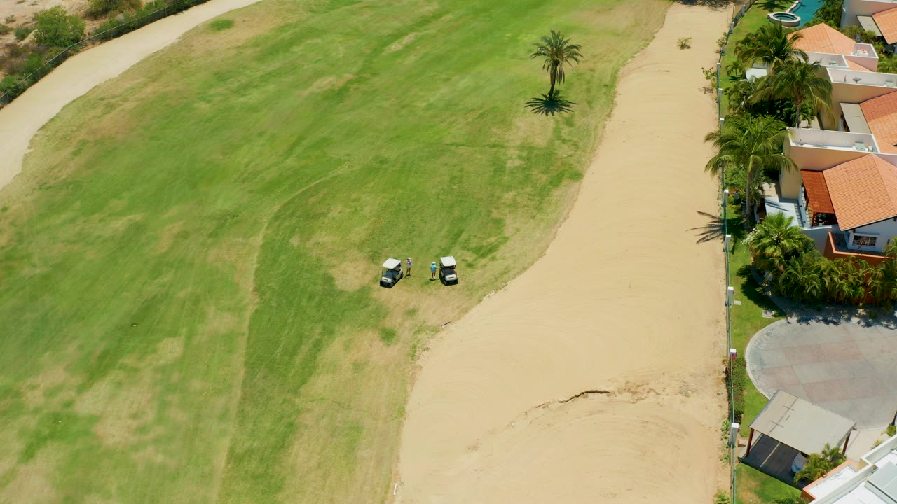 High aerial frame over a San Jose del Cabo golf course edge with two golf carts near a beige sandy path, green fairway grass, palms, red-tile roofs, and no visible people.