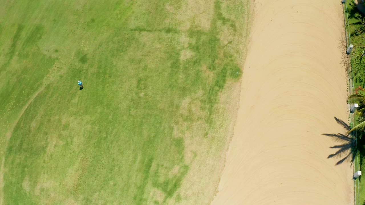 Overhead frame from San Jose Del Cabo, Baja California Sur, showing a green field, beige sandy path, palm-edge vegetation, a small vehicle near the upper-left lawn area, and no visible people.