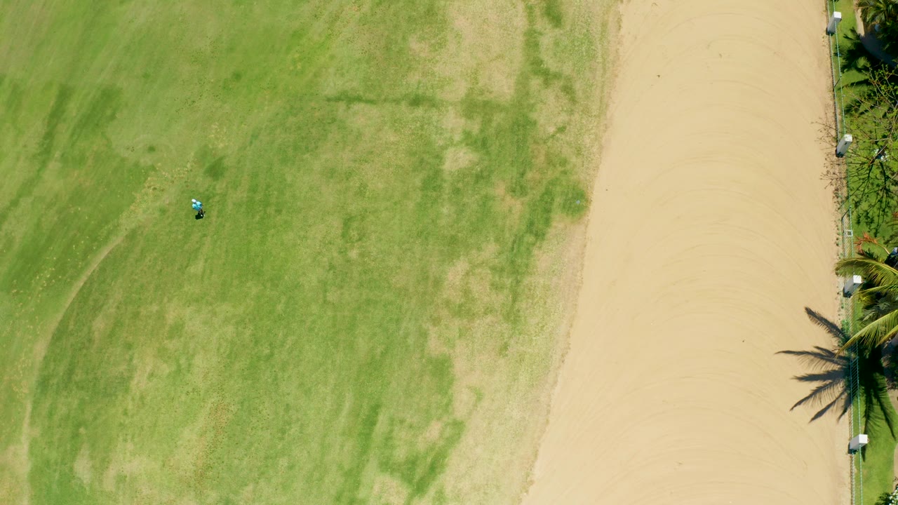 Aerial frame from San Jose Del Cabo, Baja California Sur, showing patchy green grass, a pale sandy path, a tiny vehicle on the field, palm trees at the edge, and no visible people.