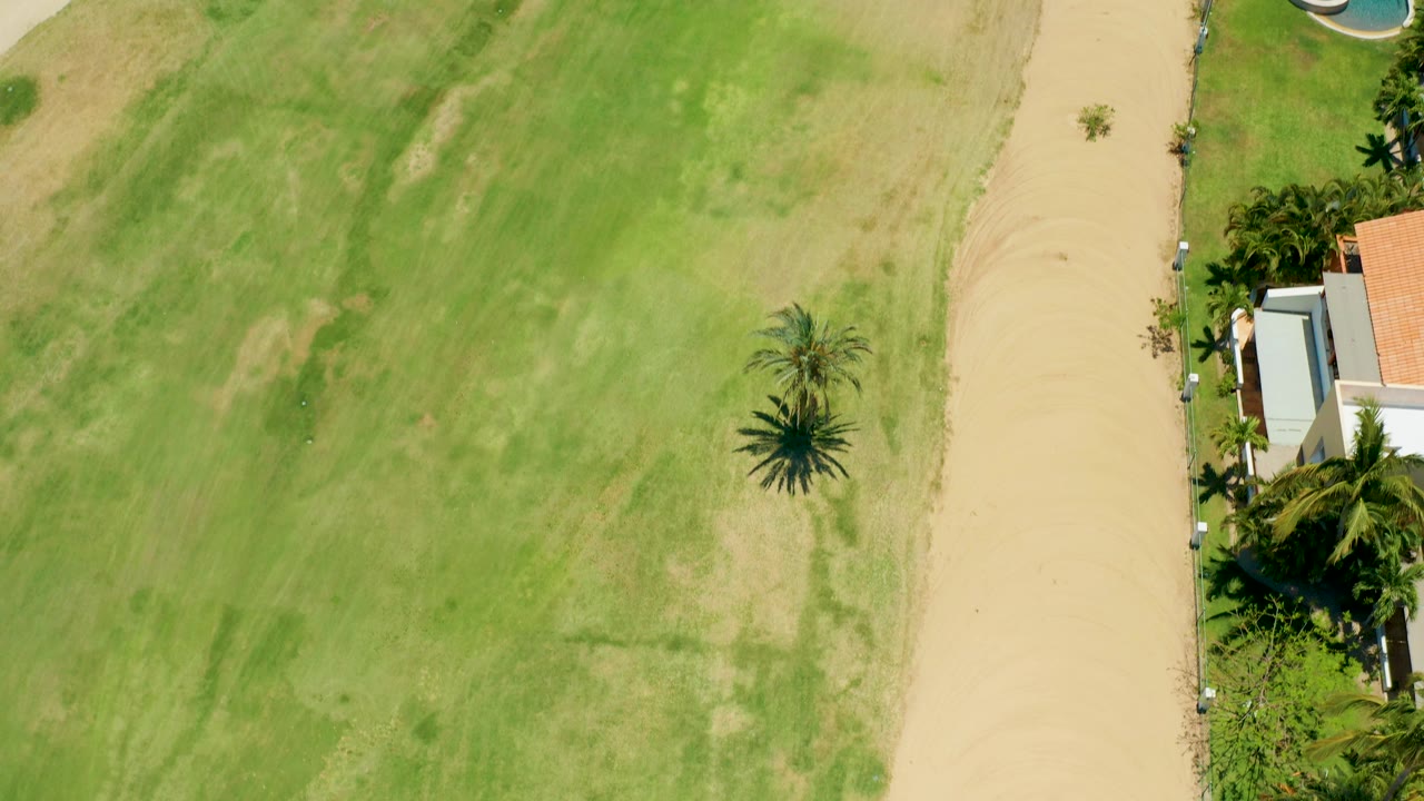 Aerial footage over San Jose Del Cabo shows more of the field, a vertical dirt path, palms, and residential roofs along the right edge, with no visible people.