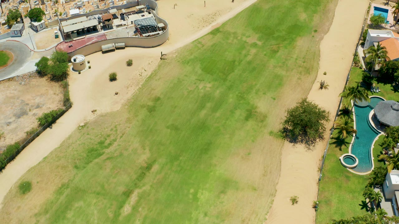 A high aerial frame over San Jose Del Cabo shows a large green field, pale sandy paths, nearby homes with pools and palm trees, and no visible people.
