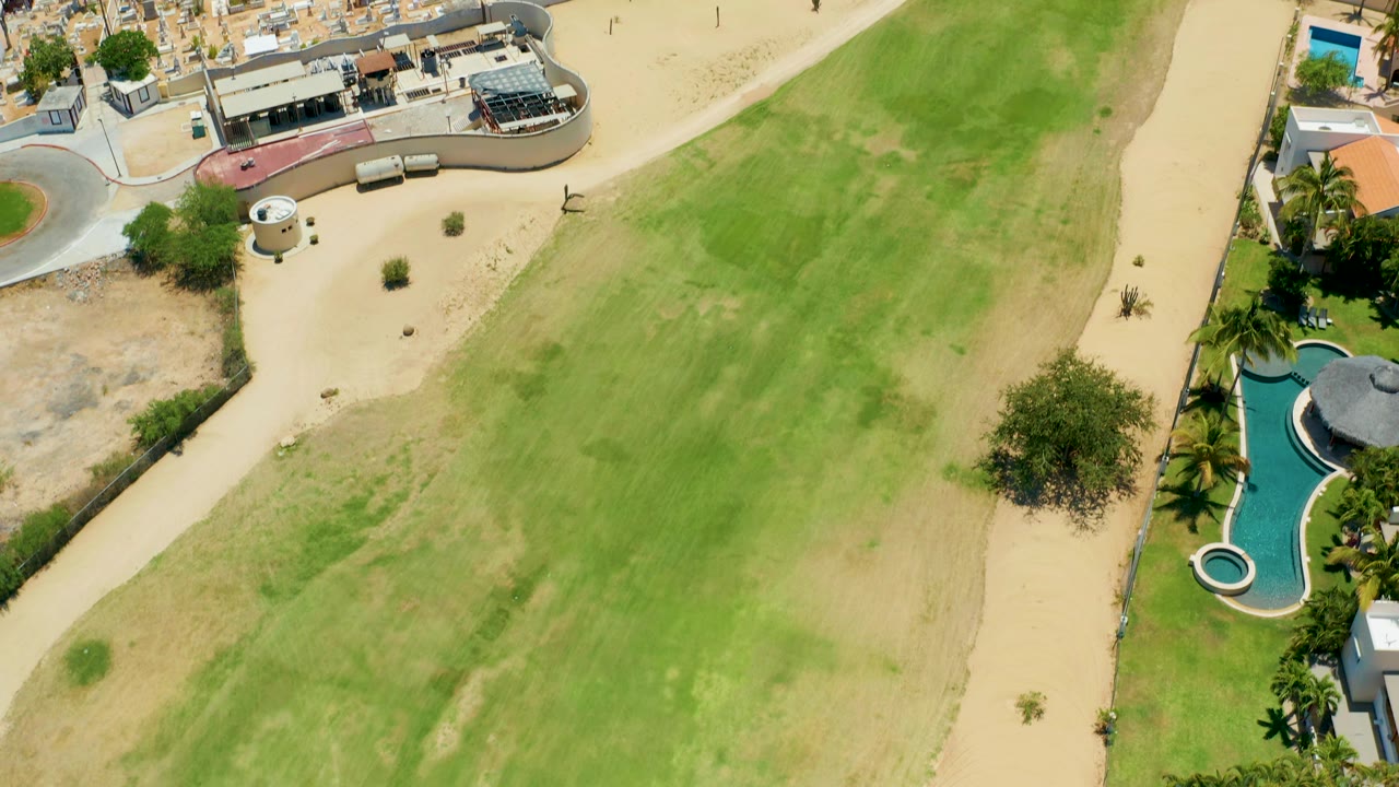 The second frame keeps the green field central while San Jose Del Cabo homes, blue pools, palms, and pale paths run along the field edge, with no visible people.