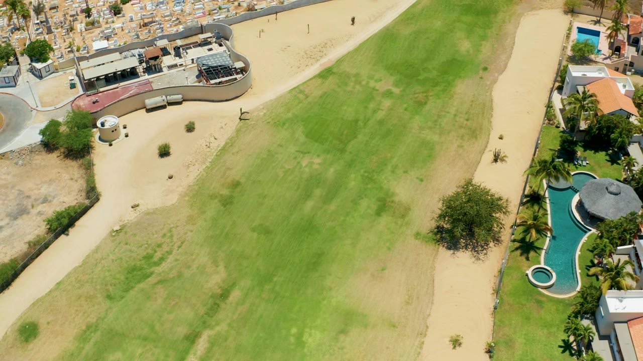 Aerial footage over San Jose Del Cabo shows the field stretching diagonally, a sandy path on one side, homes and a curved blue pool on the other, and no visible people.