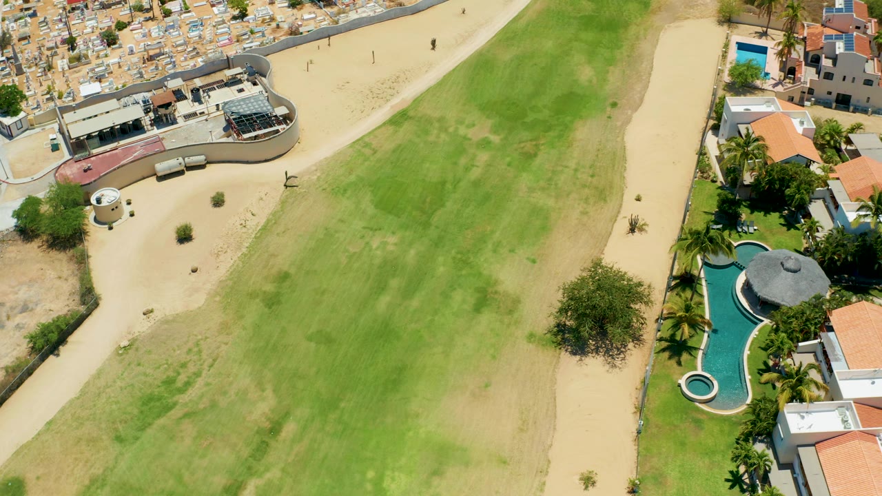 Top-down residential aerial with green grass, sandy paths, pools, roofs, and dense buildings beyond.