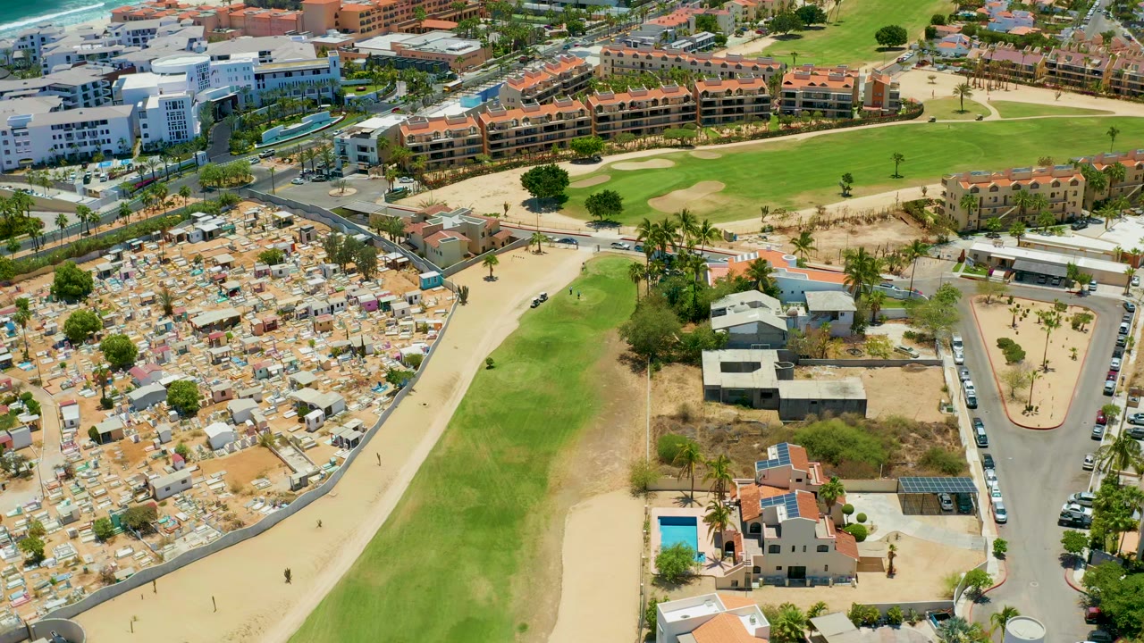 Opening aerial frame over San Jose del Cabo with crowded beach parking on the left, a long green space through the center, resort buildings beyond it, and no visible people discernible.