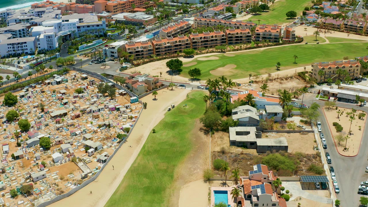 High aerial frame over San Jose del Cabo showing a curved road beside a dense tent area, bright green grass, palm trees, parked cars, and low-rise buildings, with no visible people.