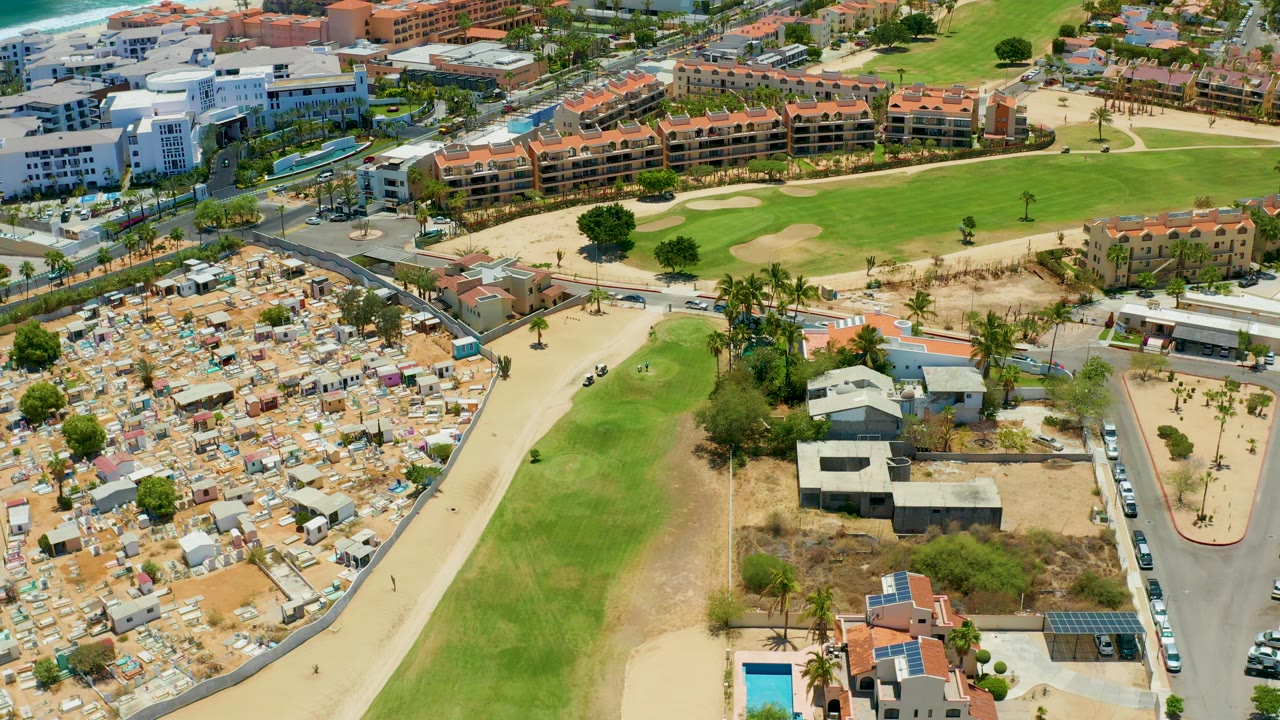 Aerial frame in San Jose del Cabo with the road and parked cars along the right side, a broad green strip in the center, tents to the left, and orange-roofed buildings in the background, with no visible people.
