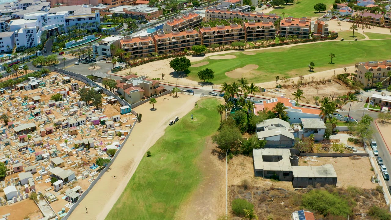 Aerial frame over San Jose del Cabo with the sandy road running between golf-course grass, compact housing, palm trees, and low buildings, with no visible people.