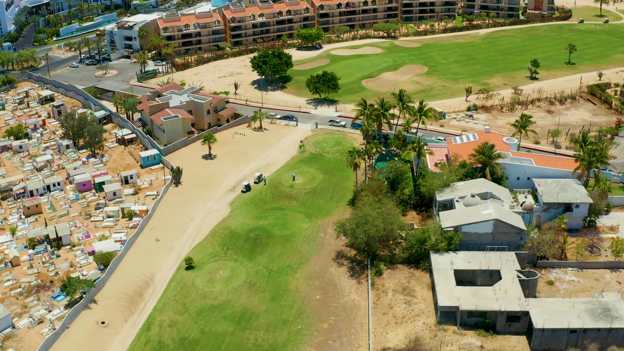 High aerial view in San Jose Del Cabo, Mexico, showing a pale cart path beside green fairways, sand traps, low buildings, palm trees, sandy lots, and no visible people.