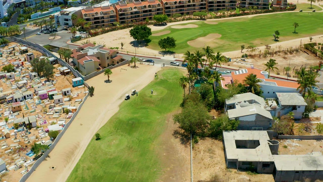 Aerial frame in San Jose Del Cabo, Mexico, with a small golf cart moving along the pale path below fairway greens, palm trees, walled homes, sandy ground, and no visible people.
