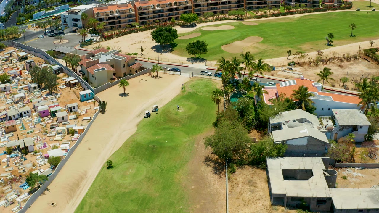 San Jose Del Cabo aerial view showing the cart path curving past bright fairway grass, tan bunkers, palms, low residential buildings, sandy lots, and no visible people.