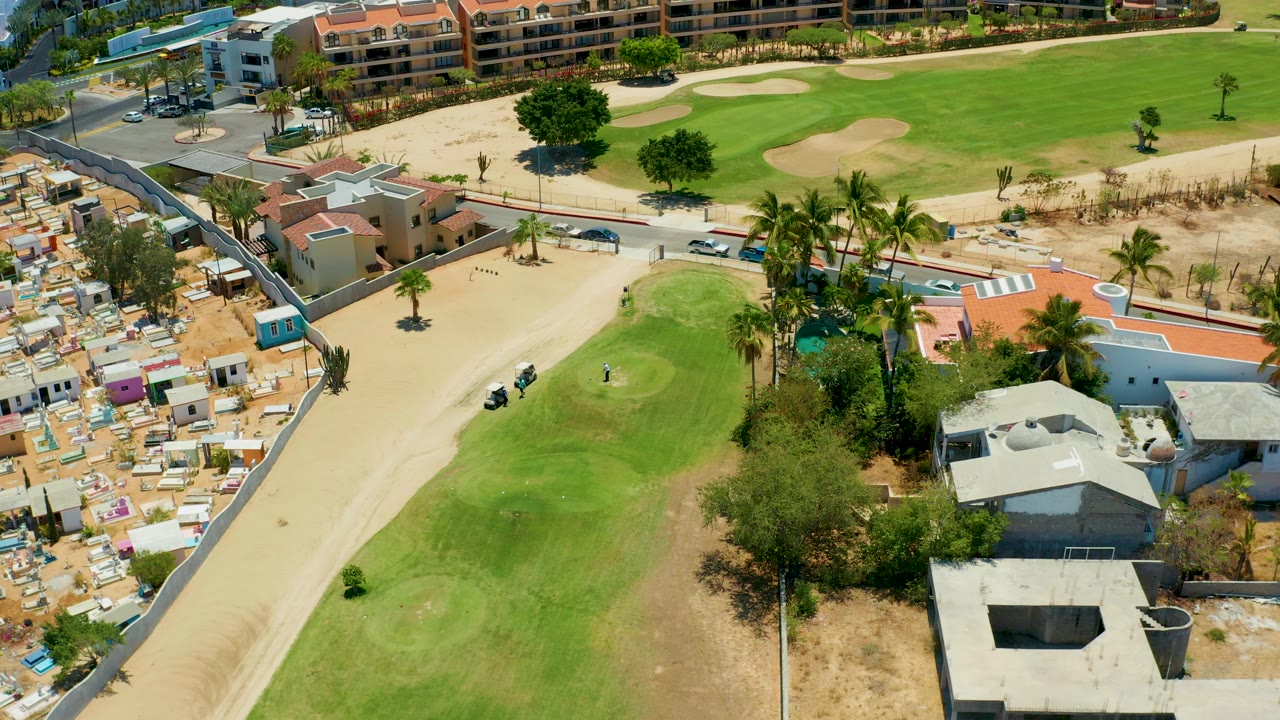 Golf cart path beside a small green, palms, sandy lots, and low buildings.