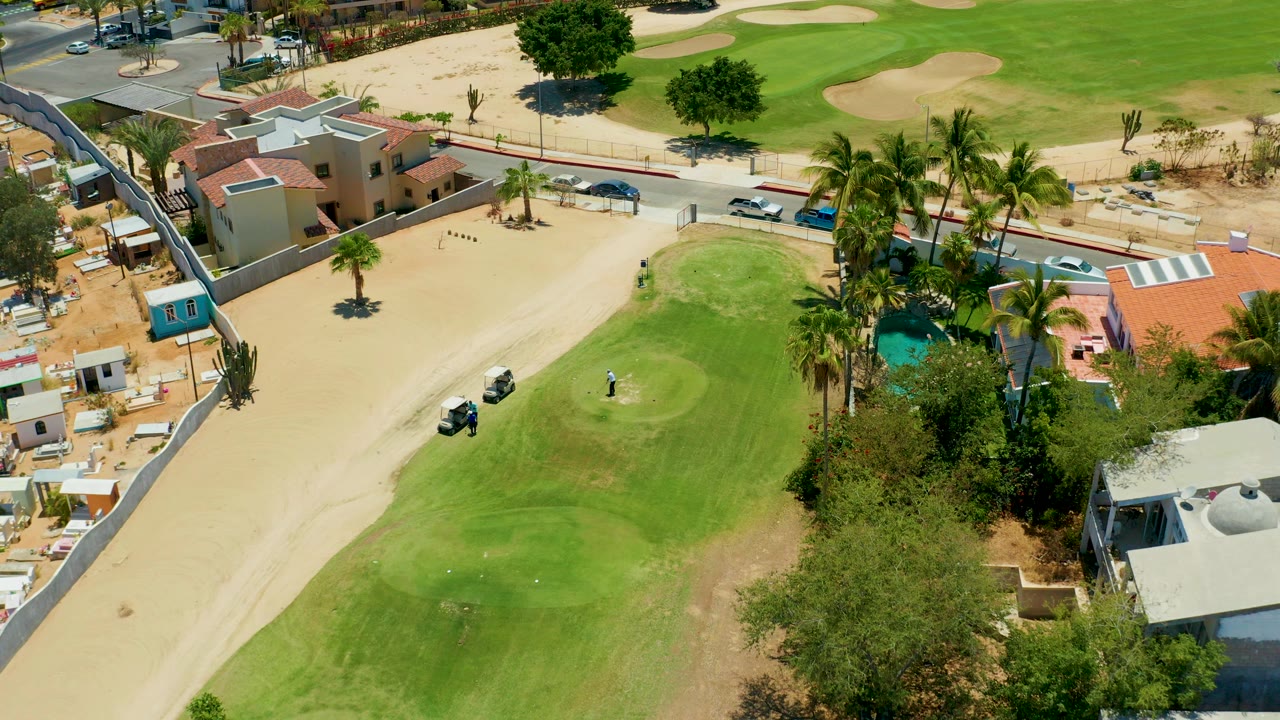 High aerial frame in San Jose Del Cabo showing a golfer on a small green, a golf cart on pale sand, surrounding homes, palms, a pool, road, and cars.
