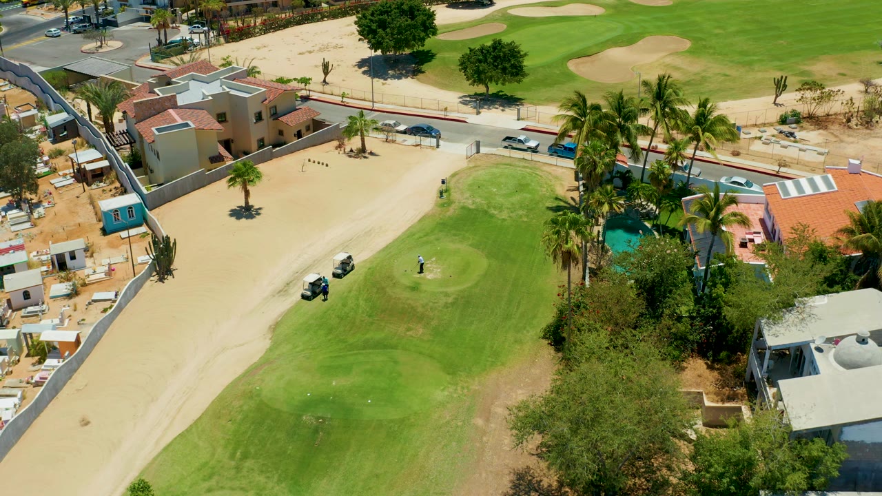 Overhead view of a golf green, sand, cart, palms, houses, pool, road, and cars.