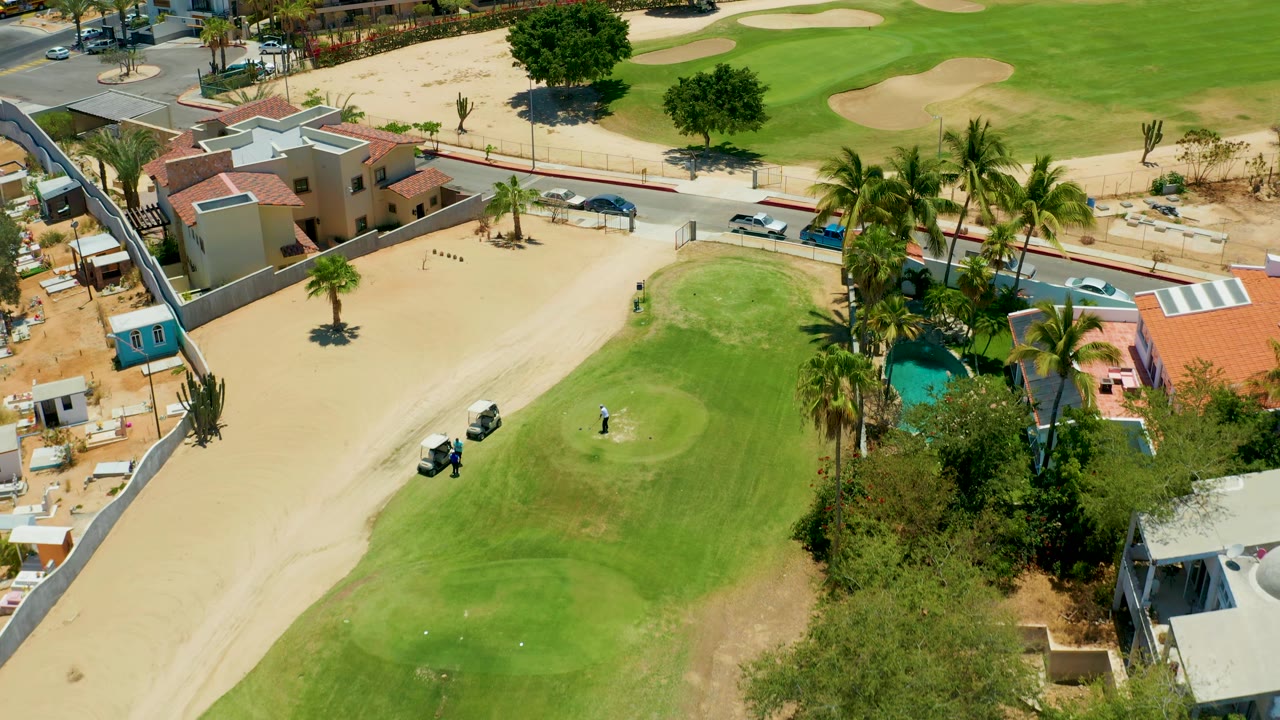 High drone frame in San Jose Del Cabo showing the golf cart lower left on sand, the golfer on the green, palms near a turquoise pool, and residential roofs near the road.