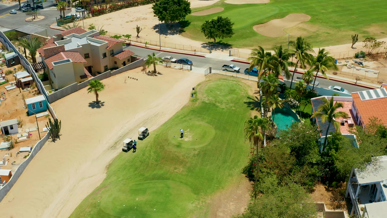 Aerial golf course frame with cart on sand, golfer on green, homes, palms, pool, road, and cars.