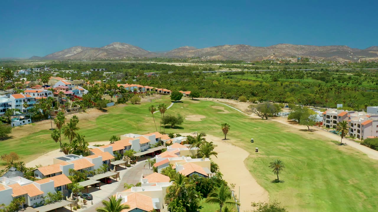 Aerial view in San Jose Del Cabo showing red-roofed homes beside a green golf course, tan sand areas, distant mountains, clear blue sky, and no visible people.