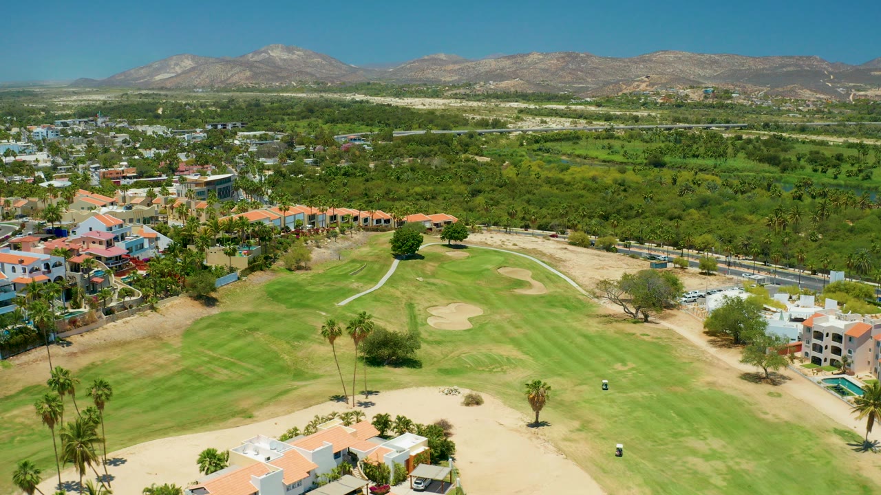 Opening aerial frame over a San Jose Del Cabo golf course with fairway grass, sand traps, red-roof homes, green vegetation, distant mountains, blue sky, and no visible people.