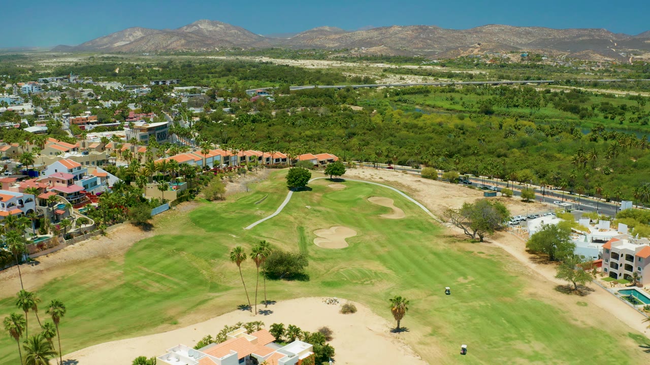 San Jose Del Cabo aerial view with a broad green golf course in front, pale bunkers, clustered red-roof houses, a road line, heavy vegetation, mountains, and no visible people.