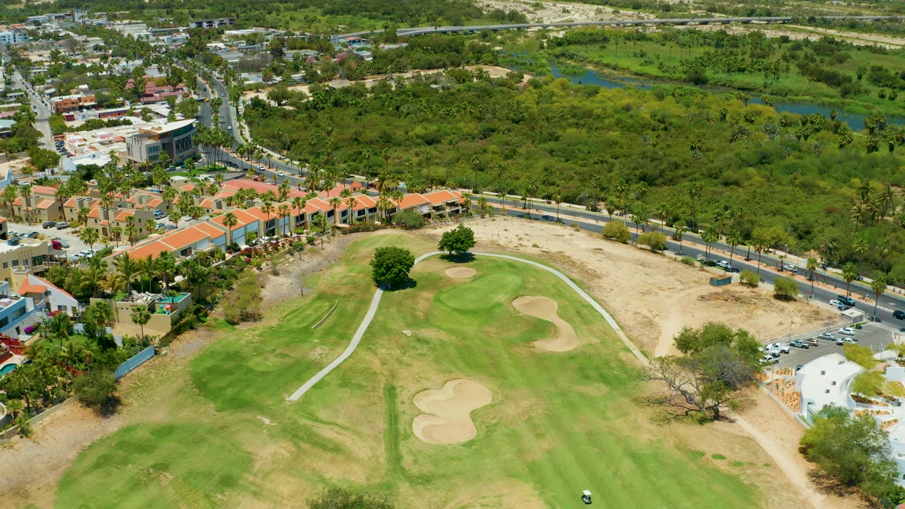 High aerial view in San Jose Del Cabo showing a green golf fairway, pale sand bunkers, orange-roofed buildings, a road with vehicles, and dense trees, with no visible people.